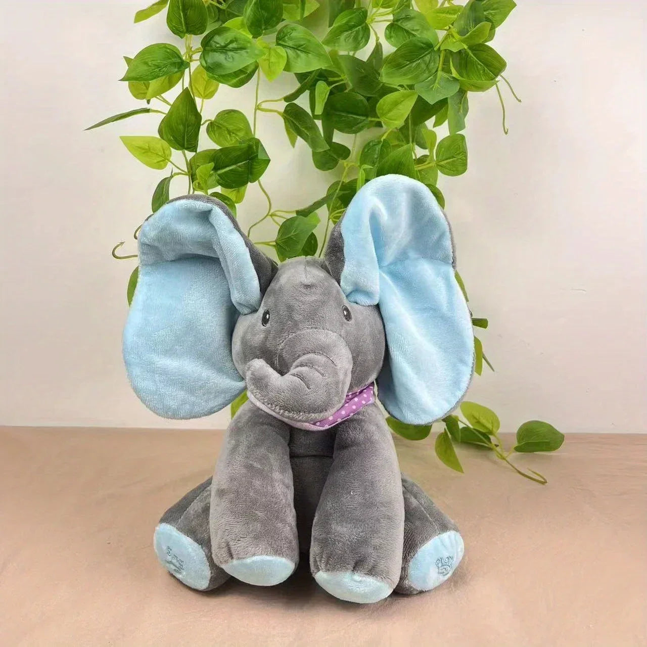 Plush elephant toy with blue ears on a beige surface with green leaves in the background