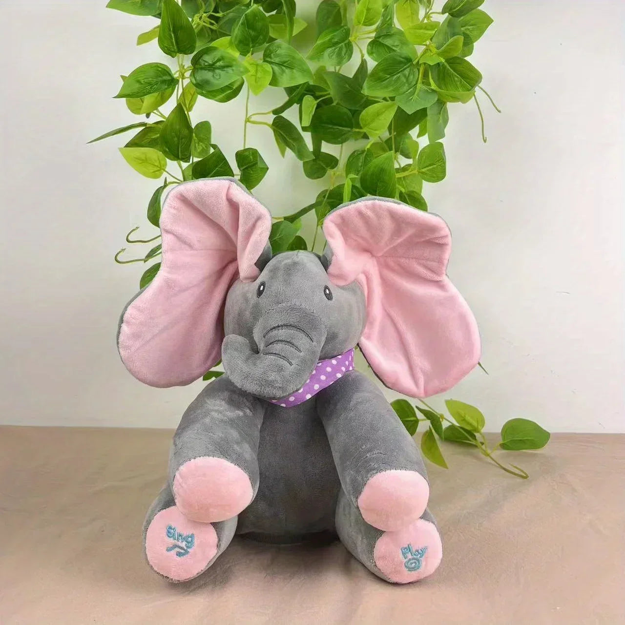 Plush elephant toy with pink ears and feet in front of green leaves